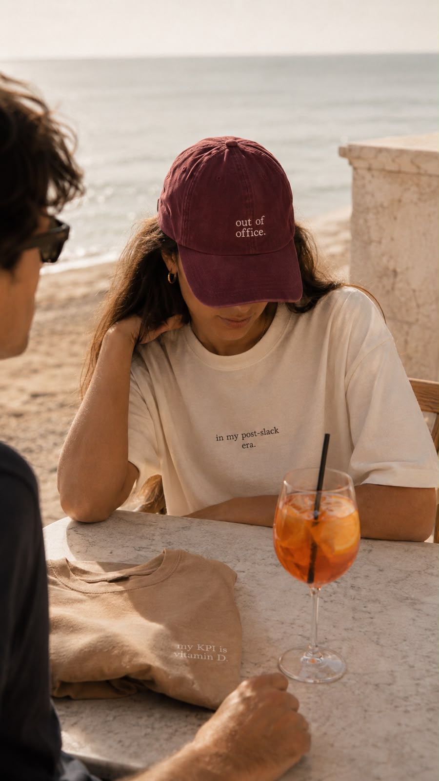 Woman at a seaside table wearing the burgundy out of office cap